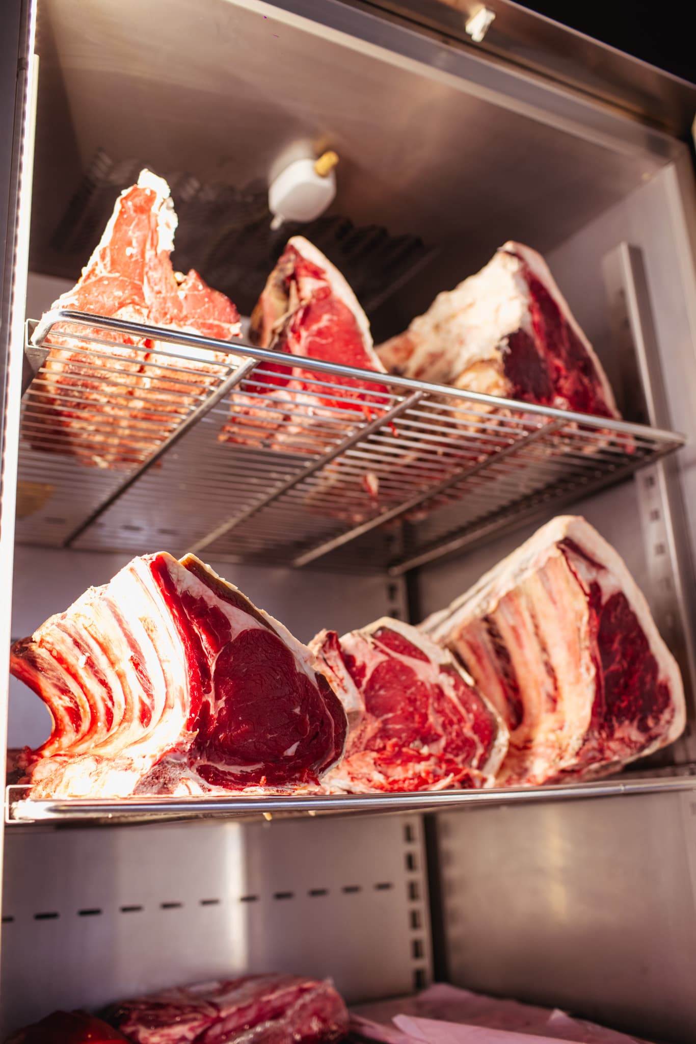 Dry-aging bone-in beef steaks, with visible marbling, displayed on metal racks in a cooler.